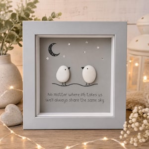 May include: A square grey frame containing pebble art of two white birds on a wire under a black crescent moon and silver stars. The text reads: No matter where life takes us we'll always share the same sky. The background is a solid grey colour.