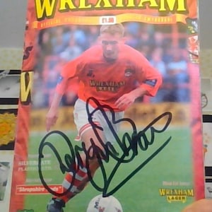 May include: A Wrexham football program cover with a player in an orange jersey, signed in black ink. The program features the text "WREXHAM" in large yellow letters. The cover also includes the text "V. SHREWSBURY TOWN" and "Tuesday, 8th October 1996 Kick-off 7.30pm."