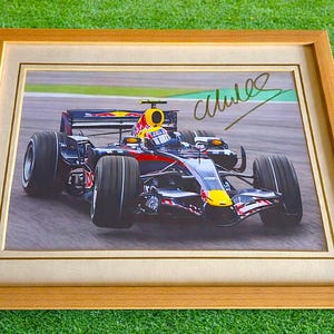 May include: Framed print of a Formula 1 race car in motion. The car is primarily black, red, and yellow, with sponsor logos visible. The print is in a wooden frame with a cream-colored mat, set against a green background.