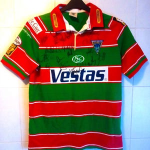 May include: A green, red, and white rugby shirt with short sleeves, displaying the text "Vestas" across the chest. The shirt has a collared neckline and is decorated with logos and signatures. The Warrington Wolves logo is visible.