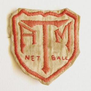 Könnte beinhalten: Beigefarbener Stoffaufnäher mit roter gestickter Umrandung und Schriftzug. Der Aufnäher zeigt die Buchstaben HTM und die Worte NET BALL. Die rote Stickerei umrandet die Form und die Buchstaben.