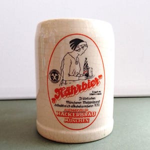 May include: A white ceramic beer stein with a red and black logo for "Nahrbier Diätetisches Münchener Malspräparat extraktreich alkoholarm (unter 1½%) ALLEINHERSTELLER HACKERBRAU MUNCHEN". The logo features a black and white illustration of a woman serving a beer.