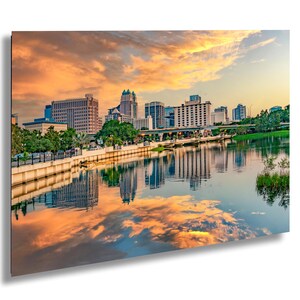 Puede incluir: Fotografía de un horizonte urbano al atardecer. La imagen presenta una masa de agua que refleja los edificios y el cielo colorido, con tonos de naranja, amarillo y azul. Los edificios son una mezcla de arquitectura moderna y clásica.