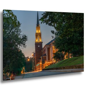 Könnte beinhalten: Ein Foto einer Kirche mit einem hohen, im Abenddämmerung beleuchteten Turm. Das Gebäude ist aus Backstein und hat eine Uhr. Bäume umrahmen die Kirche und eine Straße führt dorthin.