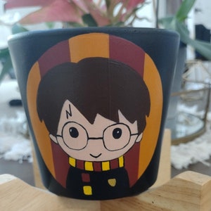 May include: A black ceramic plant pot featuring a hand-painted cartoon illustration of a boy with dark hair, round glasses, and a lightning bolt scar on his forehead. The character wears a scarf with yellow and red stripes, set against a circular background.