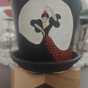 May include: Black ceramic planter with a hand-painted image of a flamenco dancer in a red and white dress. The planter is on a wooden stand.