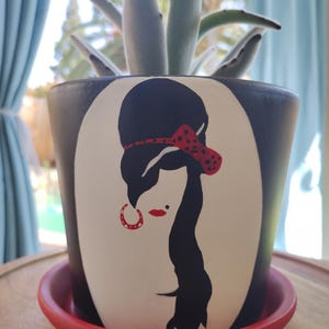 May include: A black and white painted ceramic planter with a red and white polka dot headband and large hoop earrings. The planter features a silhouette of a woman with a beehive hairstyle.