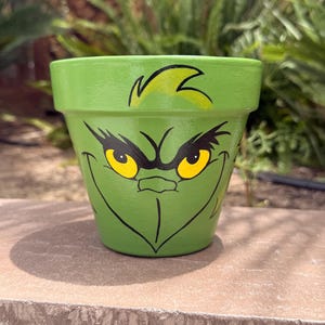 May include: A lime green terracotta plant pot featuring a hand-painted graphic of a frowning face with yellow eyes, black eyebrows, and a pointed chin. The design includes a small tuft of hair at the top rim and a wide, thin-lined mouth.