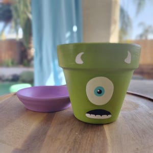 May include: A green ceramic planter pot with a painted face of a one-eyed monster. The pot has white horns and a white mouth with teeth. The pot is sitting on a wooden surface with a purple saucer.