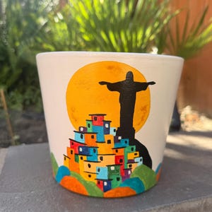 May include: A cream-colored ceramic planter featuring a hand-painted design of a black silhouette of the Christ the Redeemer statue standing before a large yellow sun, positioned above a cluster of colorful houses in shades of red, blue, and orange.