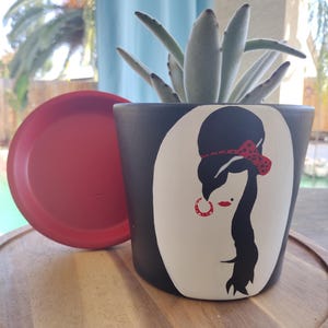 May include: A black and white ceramic planter with a hand-painted image of a woman with a red headband and earrings. The planter has a red saucer and is filled with a succulent plant.