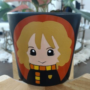 May include: A black ceramic plant pot featuring a hand-painted illustration of a character with blonde hair, large black eyes, and a small smile. The character wears a black and orange striped scarf and a black top, set against a circular orange background.