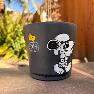May include: A dark gray cylindrical plant pot featuring a hand-painted white illustration of a cartoon dog wearing a pilot cap and goggles, accompanied by a small yellow bird character sitting inside a gray geometric shape on the side.