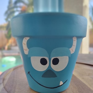 May include: A blue ceramic planter with a hand-painted cartoon face of a blue monster with white eyes, a black nose, and a white tooth.
