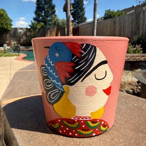 May include: A terracotta-colored flower pot featuring a painted portrait of a woman with a blue bird. The woman has black hair, a red lip, and a yellow top. The pot has a red and green patterned border.