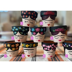 May include: A collection of hand-painted terracotta flower pots. Each pot features a stylized face with closed eyes, a red heart-shaped mouth, and a floral or patterned design around the rim. The pots are in various colors and patterns.