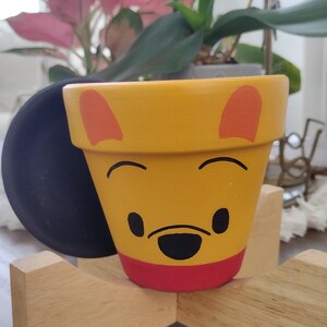 May include: A yellow ceramic planter with a painted Winnie the Pooh face. The planter has orange ears, black eyes, and a black nose. The planter is sitting on a wooden stand.