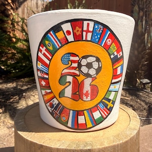 May include: A white ceramic planter featuring a hand-painted design of the number 2026 with a soccer ball. The numbers are decorated with various national flags, and the central yellow circle is surrounded by a border of many international flags.