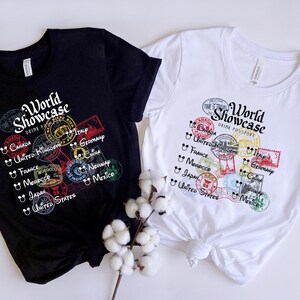 May include: Two black and white t-shirts with a graphic print of colorful stamps and the text "World Showcase Drink Passport". The stamps feature the names of countries like Canada, United Kingdom, France, Morocco, Italy, Germany, China, Norway, Mexico, and Japan.