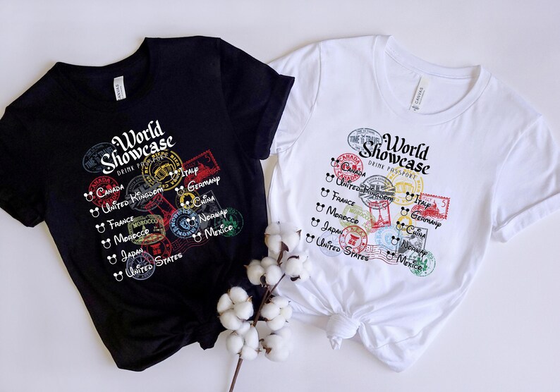 May include: Two black and white t-shirts with a graphic print of colorful stamps and the text "World Showcase Drink Passport". The stamps feature the names of countries like Canada, United Kingdom, France, Morocco, Italy, Germany, China, Norway, Mexico, and Japan.