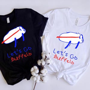 May include: Two t-shirts, one black and one white, each featuring a cartoon buffalo design with the text "Let's Go Buffalo." The buffalo is white with a red stripe and blue legs. The text is in blue and red.