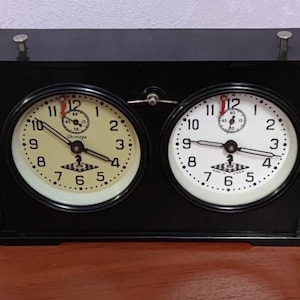 Könnte beinhalten: Eine schwarze Schach Uhr mit zwei weißen Zifferblättern, jedes mit schwarzen Zahlen und Zeigern. Die Uhr steht auf einer Holzoberfläche mit Schachfiguren in der Nähe.