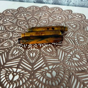 May include: Three cylindrical pen barrels arranged horizontally, each featuring a unique marbled pattern in shades of amber, brown, and black. The barrels have a smooth, polished finish with distinct threaded ends, showcasing a glossy resin-like texture.