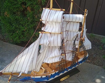 May include: A wooden model ship with white and blue sails. The ship is on a black base and has the text "ORCA NES VICTORIA" on the side.