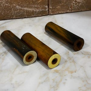 Puede incluir: Tres objetos cilíndricos con un exterior veteado de color marrón oscuro y un extremo circular liso de color crema claro que presenta un pequeño orificio centrado. Los objetos tienen un acabado liso y pulido y una forma tubular constante en toda su longitud.