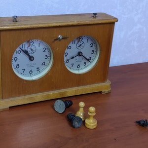 May include: A vintage wooden chess clock with two circular faces, each displaying a clock face with the text "UAC 230 MOCKBA". Chess pieces, including pawns and a king, are scattered on the table.