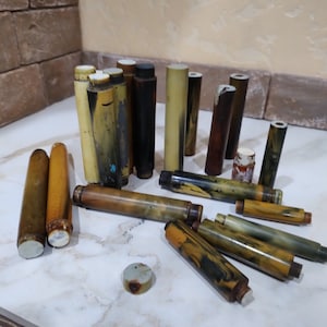 Puede incluir: Una colección de piezas cilíndricas para bolígrafos con patrones jaspeados en tonos marrón, amarillo, negro y crema. Estas piezas de resina o acrílico de diversas longitudes tienen extremos planos y muestran signos de desgaste, con un disco pequeño.