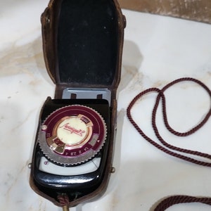 May include: A vintage brown leather case with a black and silver light meter inside. The light meter has a red and white dial with the word "Lumulight" printed on it. The case has a brown cord attached to it.