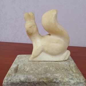 Puede incluir: Una escultura de mármol blanco de una ardilla sentada sobre una base de mármol. La ardilla está en posición sentada con la cola enroscada detrás de ella.