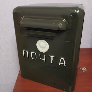 Pode incluir: Uma caixa de correio de metal verde escuro com um rótulo branco que diz "ПОЧТА" e um círculo branco com um desenho no centro. A caixa de correio tem uma fenda para o correio e uma fechadura.