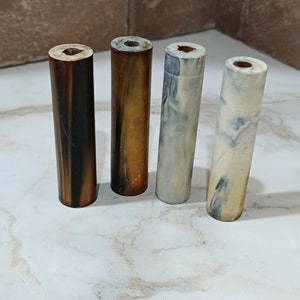 May include: Four different coloured cylindrical pen blanks. The blanks are made of a marbled material with brown, black, white, and blue colours.