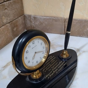 May include: A black desk set with a clock, pen holder, and pen. The clock face is white with black numbers and gold hands. The pen is black with a gold trim. The desk set has a black base with gold lettering that reads "очередный день театра-6 знак глубокие Уважения, любви и благодарности от дирекции Могилев-Подольскоговѣк и Народного театра 27 марта 1968 г."
