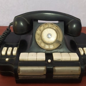 以下が含まれることがあります： 白いダイヤルと白いボタンがついた黒いロータリーダイヤル式電話。電話は黒いコードが付いており、木製の表面の上に置かれています。