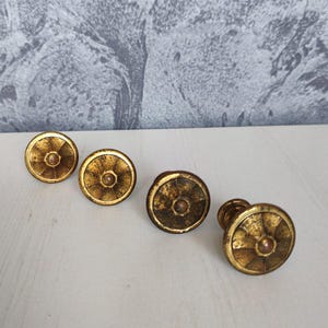 May include: Four antique brass cabinet knobs with a circular design. The knobs have a gold finish and a central, raised detail. They are arranged on a white surface against a textured gray background.