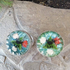 Pode incluir: Dois botões de porta redondos, prateados, com um inserto de vidro transparente com um design floral. O design floral inclui rosas vermelhas, brancas e verdes sobre um fundo azul.