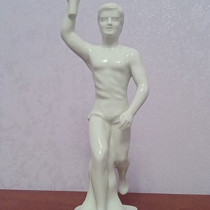 May include: White porcelain figurine of a runner holding a torch, standing on a base with the Olympic rings.