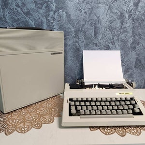 Può includere: Una macchina da scrivere vintage bianca con una tastiera nera e una custodia bianca. La macchina da scrivere è etichettata "Princesa" e si trova su una superficie bianca con un pizzo di pizzo marrone.