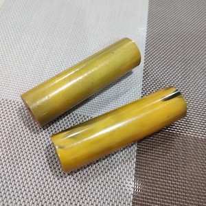 Puede incluir: Dos objetos cilíndricos con un acabado liso y pulido en tonos amarillo mostaza y ámbar. Cada pieza presenta sutiles vetas oscuras de aspecto natural y variaciones de color, con una apariencia sólida y opaca y extremos circulares planos.