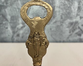 May include: A gold-colored bottle opener shaped like a bell with an ornate design featuring an elephant head.