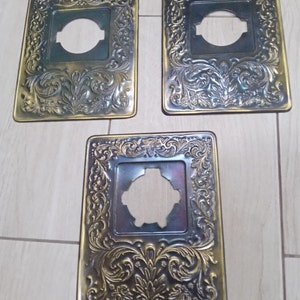 Puede incluir: Tres tapas de tomacorriente de metal adornadas con un diseño floral decorativo. Las tapas son doradas y tienen una abertura circular en el centro.