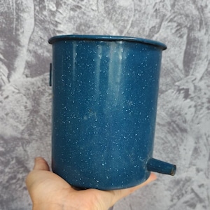May include: A blue enamelware water dispenser with white speckles. The vintage container has a spout and a handle. The cylindrical shape is held by a hand.