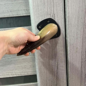 May include: A door handle with a dark brown, curved grip and a black mounting plate. The handle is held against a light gray wooden door with vertical grain. The door has a dark gray horizontal accent.