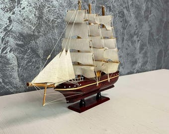 May include: A miniature wooden sailboat model with cream-colored sails and a dark red hull. The model has detailed rigging and is mounted on a dark red base. The background is a textured gray wall.