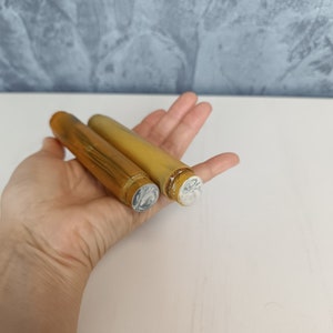 May include: Two gold-coloured metal tubes with white caps. The tubes are different sizes and are held in a hand.