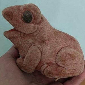 May include: A plush, light brown frog toy with a textured surface. The frog has large, dark eyes and an open mouth revealing a red interior. The toy is held in a hand.