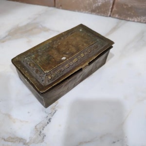 Könnte beinhalten: Eine rechteckige antike Messingbox mit Scharnierdeckel. Der Deckel und die Seiten weisen Gebrauchsspuren auf, mit dekorativen Details an den Rändern. Die Box liegt auf einer weiß-grauen Marmoroberfläche.